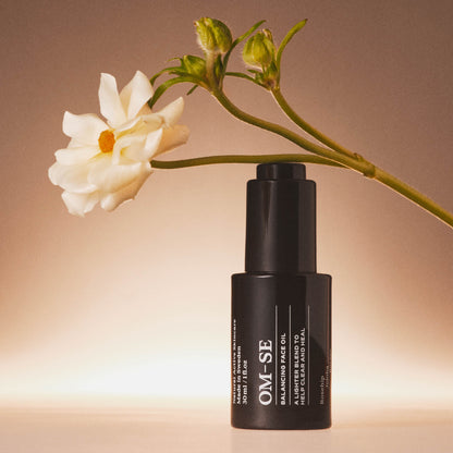 Black bottle of OM-SE Balancing Face Oil with a white flower on a gradient background