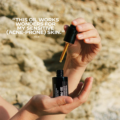 Hand holding a dropper over a bottle of OM-SE Balancing Face Oil.