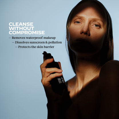 Woman holding an OM-SE Face Cleansing Oil bottle with skin benefit text on a blue background: Cleans without compromise, removes waterproof makeup, dissolves sunscreen and pollution, protects the skin barrier.