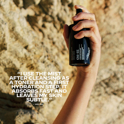Hand holding an OM-SE Hydrating Face Mist bottle with customer text overlay on a textured stone background.