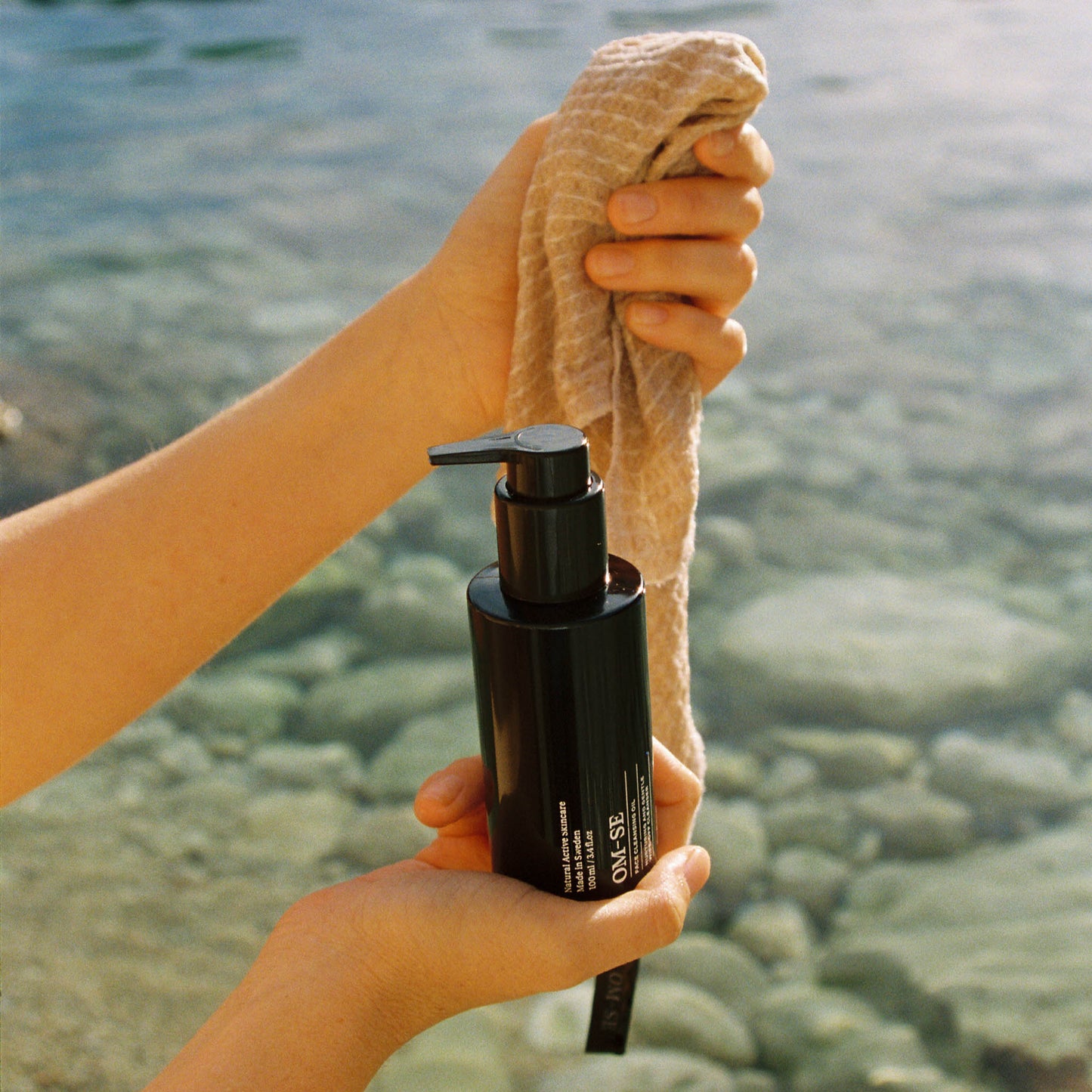 Hand holding a bottle of OM-SE Face Cleansing Oil and a linen face cloth